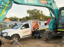Man standing beside an excavator and Orange Hire vehicle at a construction site with safety barriers in place.